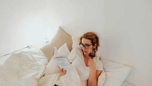 A postmenopausal woman reads a book and holds a glass of wine while relaxing in bed.